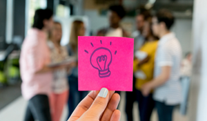 A picture of a woman's and holding a pink post it note with a light bulb drawn on it.