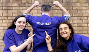 A picture of two women, smiling and one male facing backwards, wearing purple t-shirts, with the words volunteer wrote on the t-shirts.