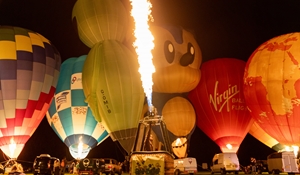 Hot air Balloons tethered on a field. They have their gas burners lit and there are people and vans around them. It is night time