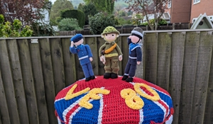 A VE Day celebration knitted post-box topper showing three services in uniform, standing on the union flag