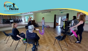 Group exercise session with older adults seated in chairs, following an instructor at the front of the room holding colourful scarves. The move to thrive logo is displayed at the top