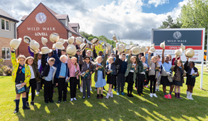 A group of school children in uniform standing on a lawn, holding up safari hats in celebration in front of a housing sign.