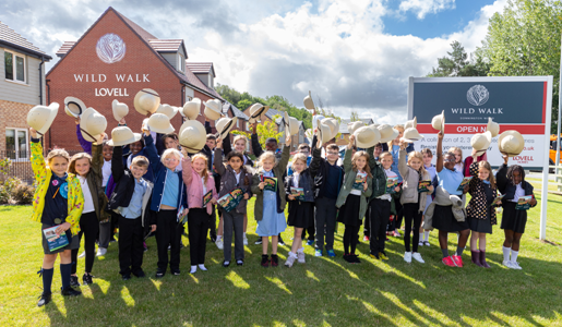 A group of school children in uniform standing on a lawn, holding up safari hats in celebration in front of a housing sign.