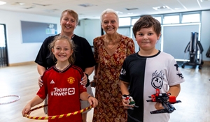 A group of two children and two adults smiling in a community room; the girl holds a hula hoop and the boy holds a toy.