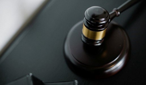 A dark wooden judge's gavel with a gold band resting on a circular sounding block against a dark background.